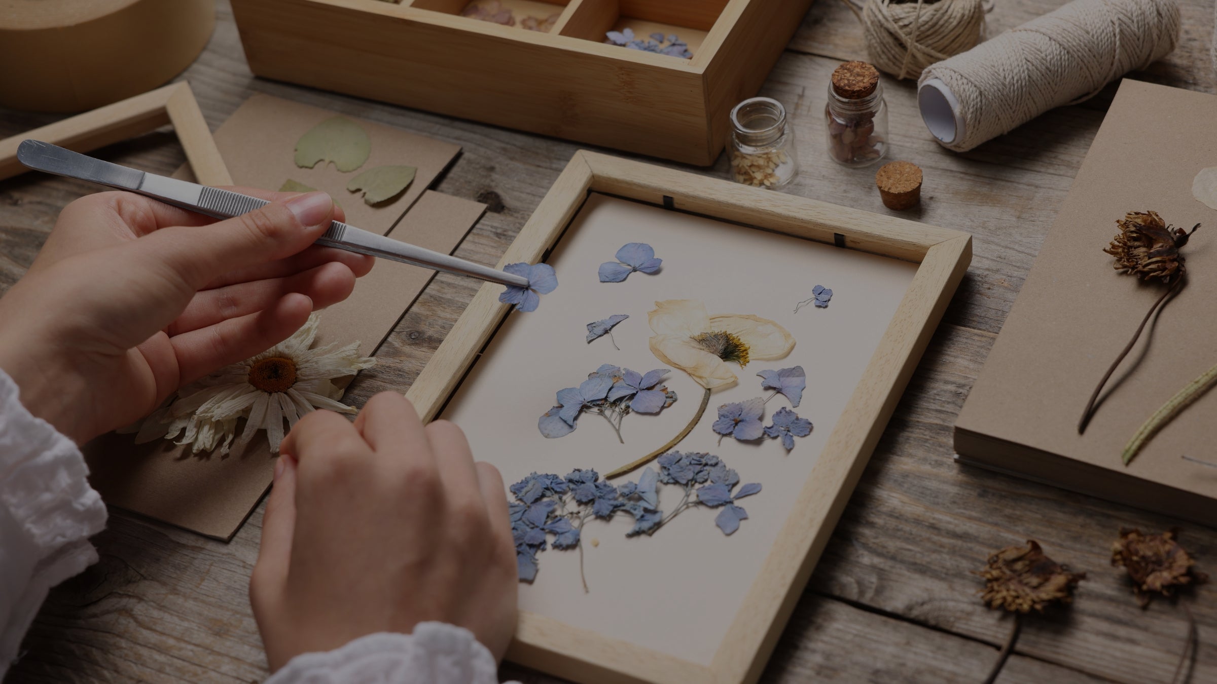 Person arranging dried flowers in a wooden frame on a wooden table.