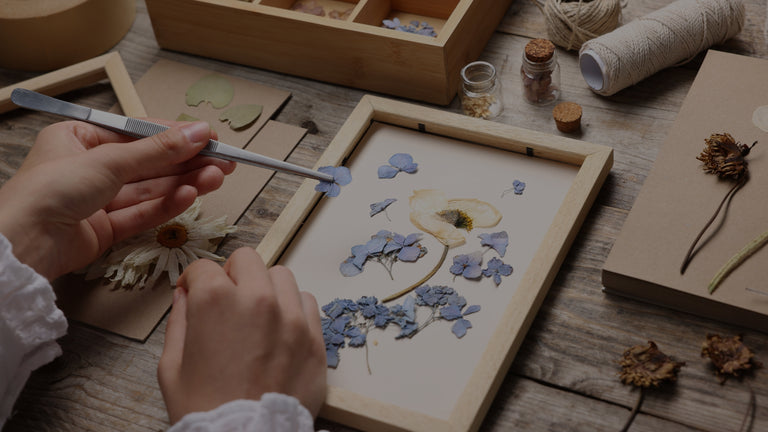 Person arranging dried flowers in a wooden frame on a wooden table.
